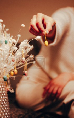 A woman hangs a yellow decorative Easter egg on a bouquet of dry white flowers in the interior. Festive home decor. Woman's hand holding toy pendant. 