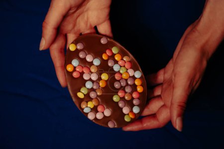 Large chocolate Easter egg with colorful candies in female hands Dark table top view. Easter's treats and sweets. Festive food background. Egg hunting