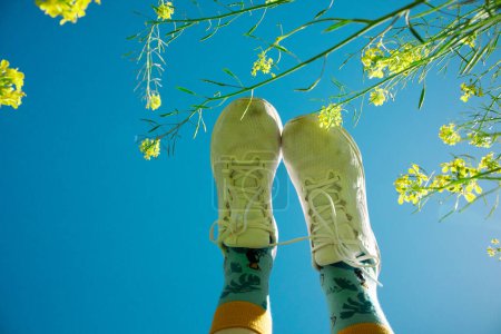 Woman's feet in white sneakers raised up against the blue sky. A girl resting on a spring meadow among blooming Yellow mustard flowers in spring day. 