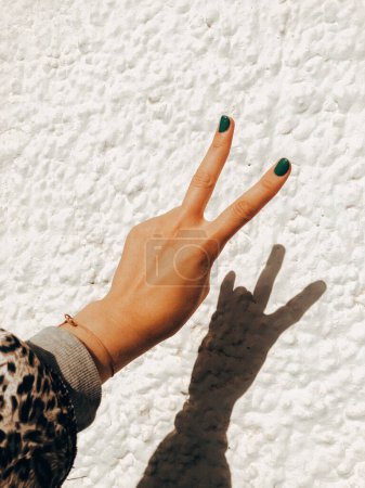 Photo for Female hand makes a two finger up greeting gesture on white stone wall background. Green manicure. Hipster woman. Good mood, peace victory sign. - Royalty Free Image