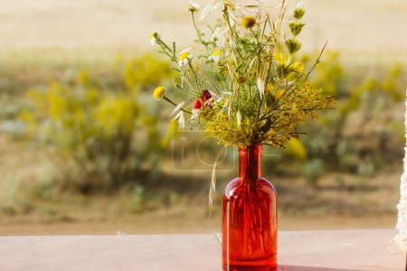 Bouquet of fresh wildflowers in a red glass vase on the window sill against the background of a field, meadow on a sunny day. Modern decor for rural home. Bunch of flowers and plants in sunny day.