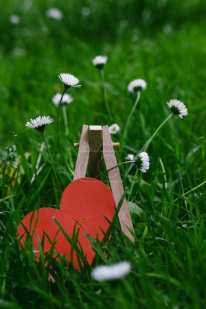 Red paper heart on lawn with green grass and white daisy flowers. Falling in love, love, feelings concept. Vintage pale colors, cinematic effect. 