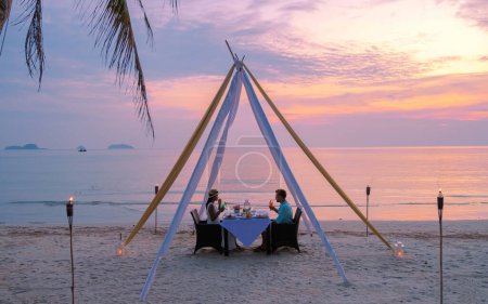 Photo for A couple of men and women having a Romantic dinner on a beach in Koh Chang Thailand during sunset. Men and women with Table with dinner on a beach - Royalty Free Image