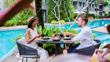 Téléchargez les photos : Couple prenant le petit déjeuner au bord de la piscine pendant les vacances. Asiatiques femmes thaïlandaises et hommes caucasiens prendre le petit déjeuner le matin - en image libre de droit