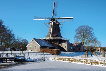 Foto de Viejo molino de viento histórico de madera en la nieve en los Países Bajos durante el invierno. Clima frío en Holanda con nieve y molino de viento en Drenthe - Imagen libre de derechos