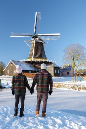 Foto de Un par de hombres y mujeres caminando en la nieve durante un frío día de invierno en los Países Bajos, con al fondo un antiguo molino de viento histórico de madera - Imagen libre de derechos
