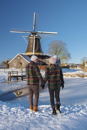 Téléchargez les photos : Un couple d'hommes et de femmes marchant dans la neige pendant une froide journée d'hiver aux Pays-Bas, avec en arrière-plan un vieux moulin à vent historique en bois dans la neige - en image libre de droit