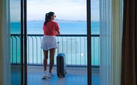 Photo for A Thai woman with hand luggage and a trolley checking in at a hotel room looking out over the ocean in Thailand. - Royalty Free Image