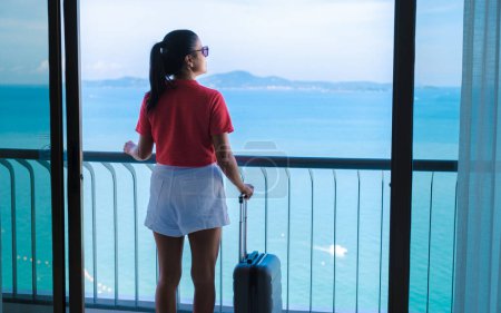 Photo for Thai women with hand luggage and a trolley checking in at a hotel room looking out over the ocean in Thailand. - Royalty Free Image