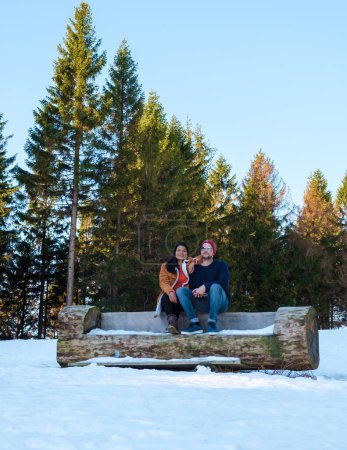 Téléchargez les photos : Couple assis sur un banc en bois dans la neige, hommes caucasiens et femmes asiatiques dans la neige avec du bois en arrière-plan au parc national du Harz en Allemagne. - en image libre de droit