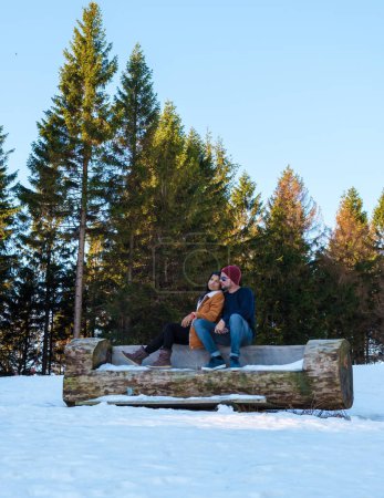 Foto de Pareja sentada en un banco de madera en la nieve, hombres caucásicos y mujeres asiáticas en la nieve con madera al fondo en el Parque Nacional Harz en Alemania. - Imagen libre de derechos