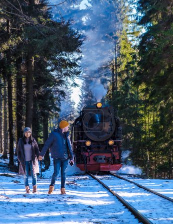Téléchargez les photos : Un couple d'hommes et de femmes regardant le train à vapeur pendant l'hiver dans la neige dans le parc national du Harz Allemagne, train à vapeur Brocken Bahn sur le chemin à travers le paysage hivernal - en image libre de droit