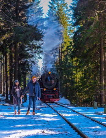 Téléchargez les photos : Un couple d'hommes caucasiens et de femmes asiatiques regardant le train à vapeur pendant l'hiver dans la neige dans le parc national du Harz Allemagne, train à vapeur Brocken Bahn à la montagne Brocken en hiver - en image libre de droit