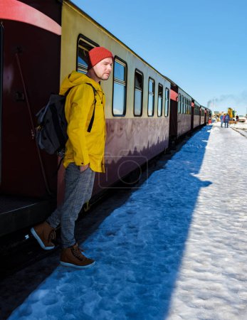 Téléchargez les photos : Jeunes hommes en imperméable au train à vapeur pendant l'hiver dans la neige dans le parc national du Harz Allemagne, train à vapeur Brocken Bahn sur le chemin à travers le paysage hivernal, Brocken, Harz Allemagne - en image libre de droit