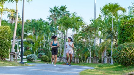 Téléchargez les photos : Deux femmes asiatiques font de l'exercice dans un parc public à Pattaya en Thaïlande. Des amies font du jogging. Femme mûre thaïlandaise exercice dans un parc avec des palmiers dans la ville - en image libre de droit