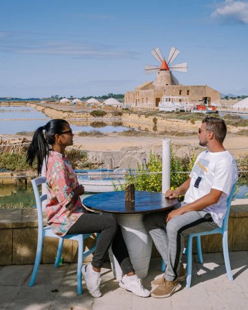 Foto de Pareja tomando café con vistas al molino de viento y a las salinas cerca de Marsala, Sicilia, Italia, hombres y mujeres de vacaciones en Sicilia - Imagen libre de derechos