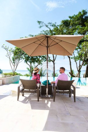 Foto de Couple man and woman mid age at a luxury beach chair, on a luxury vacation in Thailand, men and an Asian woman in a pool looking out over the bay watching the sunset. Infinity pool over the ocean - Imagen libre de derechos