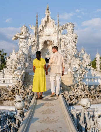 Foto de A couple visits the White Temple Chiang Rai Thailand, Wat Rong Khun, aka The White Temple, in Chiang Rai, Thailand. Asian women and Caucasian men visit a temple with a blue sky - Imagen libre de derechos