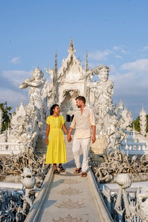 Foto de A couple visits the White Temple Chiang Rai Thailand, Wat Rong Khun, aka The White Temple, in Chiang Rai, Thailand. Asian women and Caucasian men visit a temple with a blue sky - Imagen libre de derechos