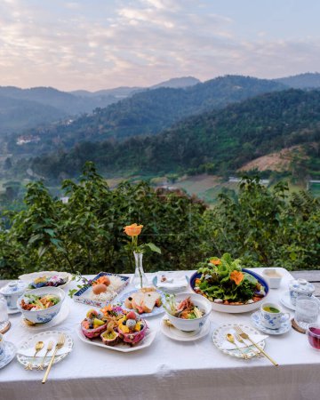 Téléchargez les photos : Breakfast table in the mountains of Thailand Chiang Mai, a wooden served festive table with homemade food and drinks, fresh fruits, and flowers under a pine tree on a sunny day. Luxury breakfast. - en image libre de droit