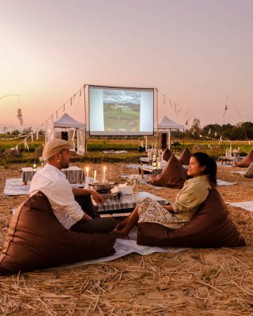 Foto de A couple of men and women watching a movie at an outdoor cinema in Northern Thailand Nan Province out over the rice paddies in Thailand, green rice fields. - Imagen libre de derechos