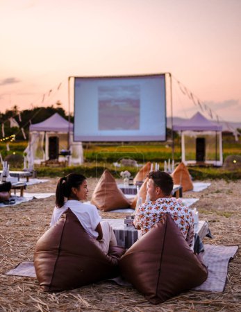 Téléchargez les photos : A couple of men and women watching a movie at an outdoor cinema in Northern Thailand Nan Province out over the rice paddies in Thailand, green rice fields. - en image libre de droit