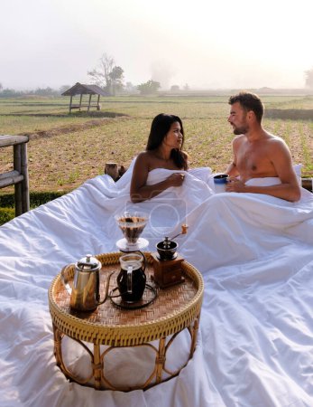 Photo for A couple of men and women at a cottage homestay in an outdoor bed in Northern Thailand Nan Province, couple waking up during sunrise in the rice fields in the morning drinking coffee outside - Royalty Free Image
