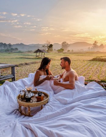 Téléchargez les photos : A couple of men and women at a cottage homestay in an outdoor bed in Northern Thailand Nan Province, couple waking up during sunrise in the rice fields in the morning drinking coffee outside - en image libre de droit