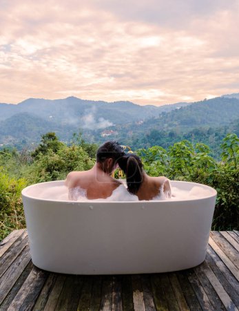 Foto de A couple of caucasian men and Asian women in a bathtub in the evening during sunset, a couple in a bathtub. - Imagen libre de derechos