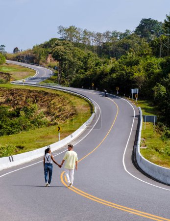 Foto de A couple walking on a curved road in the mountains of Nan Thailand, Road nr 3country road rear view. Number three road among the mountains, a couple man and women on vacation in Nan Thailand. - Imagen libre de derechos