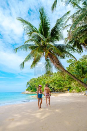 Foto de A couple of men and women walking on a white tropical beach with palm trees in Phuket Thailand. Surin Beach phuket - Imagen libre de derechos