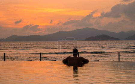 Foto de Young men at a swimming pool at a luxury hotel, a Luxury swimming pool in a tropical resort, relaxing holidays. A young man during sunset by swim pool, men watching a sunset in an infinity pool - Imagen libre de derechos
