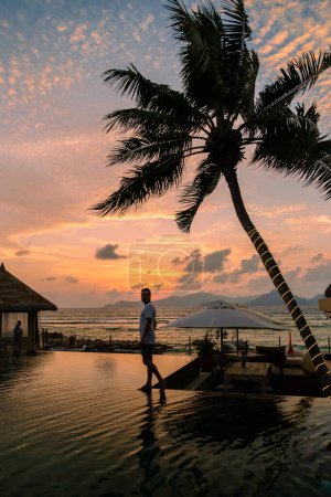 Téléchargez les photos : Young men at a swimming pool at a luxury hotel, a Luxury swimming pool in a tropical resort, relaxing holidays. A young man during sunset by swim pool, men watching a sunset in an infinity pool - en image libre de droit