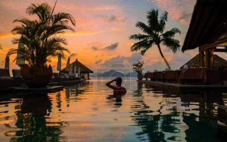 Foto de Young men at a swimming pool at a luxury hotel during sunset, a Luxury swimming pool in a tropical resort, A young man during sunset by swim pool, men watching a sunset in an infinity pool - Imagen libre de derechos
