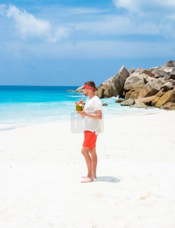 Photo for Young men in swim short with a coconut drink on a tropical beach La Digue Seychelles Islands. - Royalty Free Image