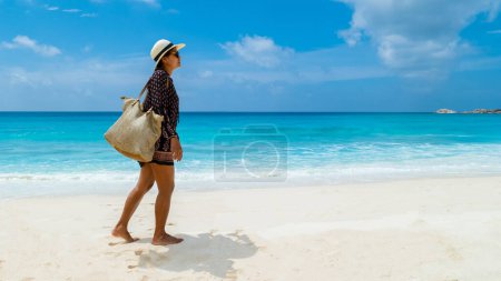 Foto de Young Asian woman with a hat walking at a tropical beach, Grand Anse beach La Digue Seychelles Islands. - Imagen libre de derechos