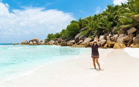 Foto de Young Asian woman with a hat walking at a tropical beach, Grand Anse beach La Digue Seychelles Islands. - Imagen libre de derechos
