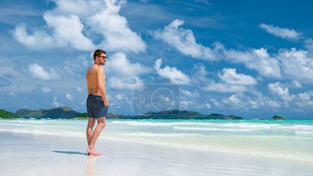 Foto de Los hombres jóvenes en nadar corto en una playa tropical blanca con el océano de color turquesa Anse Volbert playa Praslin Seychelles. - Imagen libre de derechos