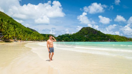 Téléchargez les photos : Young tanning men in swim short at a white tropical beach with turquoise colored ocean Anse Volbert beach Praslin Seychelles. - en image libre de droit