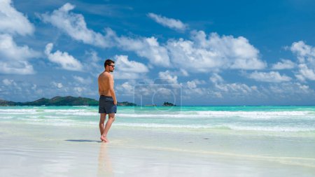 Téléchargez les photos : Jeune homme en short de bain sur une plage tropicale blanche avec un océan turquoise Anse Volbert plage Praslin Seychelles. - en image libre de droit