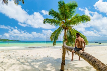 young men sitting at a palm tree on a white tropical beach with turquoise colored ocean Anse Volbert beach Praslin Seychelles.
