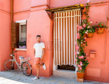 Foto de Hombres jóvenes de pie contra una pared rosa en las coloridas calles del pueblo de Burano Venecia Italia colorido canal whit barcos y casas vibrantes. - Imagen libre de derechos