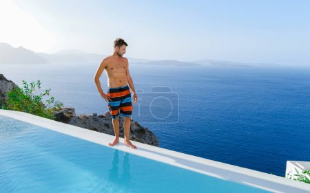 Téléchargez les photos : Young man relaxing in infinity swimming during vacation at Santorini, swimming pool looking out over the Caldera ocean of Santorini, Oia Greece, Greek Island Aegean Cyclades luxury vacation. - en image libre de droit