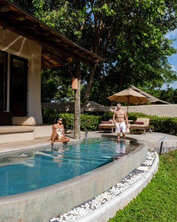 Téléchargez les photos : Un jeune couple d'hommes et de femmes dans une piscine pendant des vacances sur une île tropicale. homme et femme dans la piscine à débordement au coucher du soleil. vacances de luxe dans une villa piscine de luxe - en image libre de droit