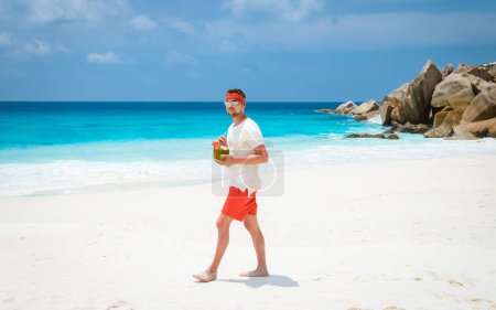 Foto de Young men in swim short with a coconut drink on a tropical beach La Digue Seychelles Islands. - Imagen libre de derechos