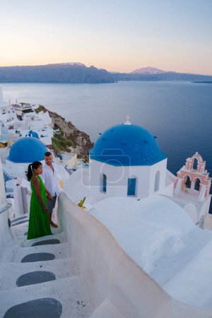 Foto de Visita de pareja Santorini Grecia, hombres y mujeres visitan el pueblo griego encalado de Oia durante las vacaciones de verano al amanecer sobre las cúpulas azules y las iglesias blancas - Imagen libre de derechos