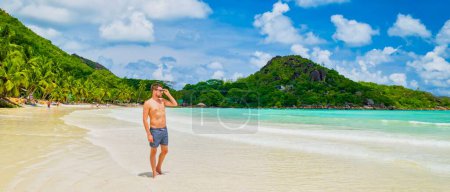 Foto de Young tanning men in swim short at a white tropical beach with turquoise colored ocean Anse Volbert beach Praslin Seychelles. - Imagen libre de derechos