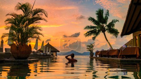Téléchargez les photos : Young men at a swimming pool at a luxury hotel, a Luxury swimming pool in a tropical resort, relaxing holidays. A young man during sunset by swim pool, men watching a sunset in an infinity pool - en image libre de droit