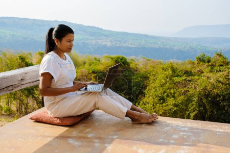 Photo for Young Asian woman digital nomad traveler working online using a laptop and enjoying the beautiful natural landscape in front of a tent at sunrise. Digital nomad working on a laptop, remote working - Royalty Free Image