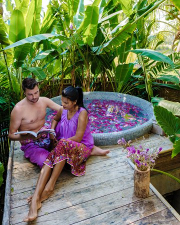 Téléchargez les photos : Couple dans une baignoire dans la forêt tropicale de Thaïlande pendant des vacances avec des fleurs dans le bain, les hommes et les femmes se détendre dans un jacuzzi avec des pétales de rose à l'extérieur dans la nature. jeunes hommes lisant un livre - en image libre de droit